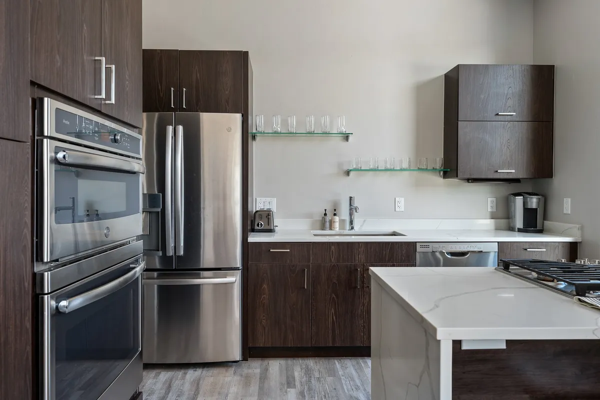A sleek, contemporary kitchen with stainless steel appliances, quartz countertops, dark wood cabinetry, and a glass shelf lined with glasses. The kitchen is ready for both everyday family meals and special gatherings, exuding a sense of culinary possibility and comfort.