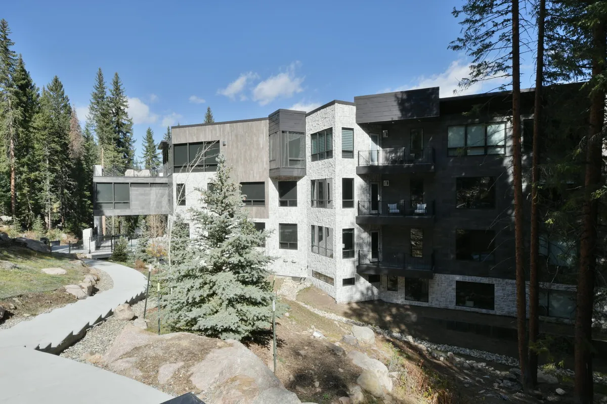 A striking view of modern architecture nestled among tall trees, with a stone pathway leading to the building's entrance. Balconies are visible, inviting guests to relax while taking in the serene forest surroundings. The peaceful scene evokes the experience of enjoying mountain views and the privacy of a luxury condo.
