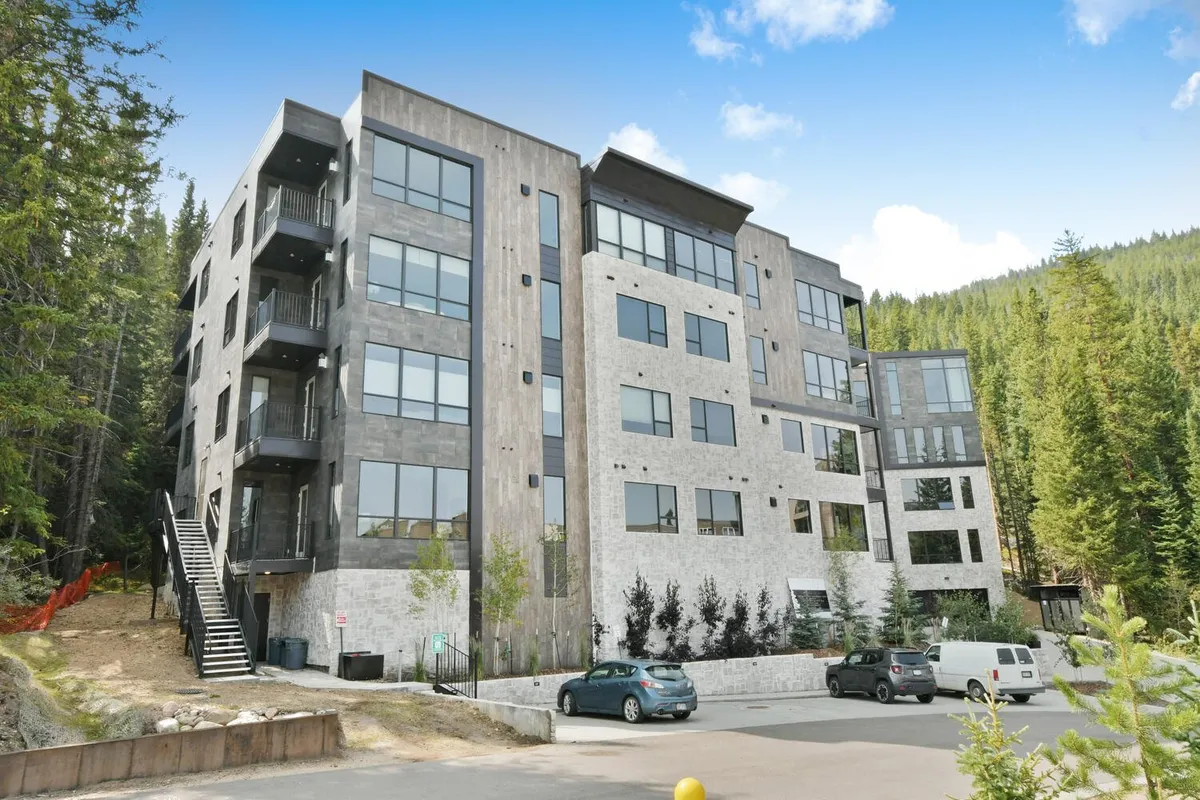 Showcases the modern architectural exterior of the building, with large windows and a wood-and-stone facade, nestled among lush greenery. The setting exudes a contemporary mountain vibe and hints at the inviting patios and balconies available to residents.