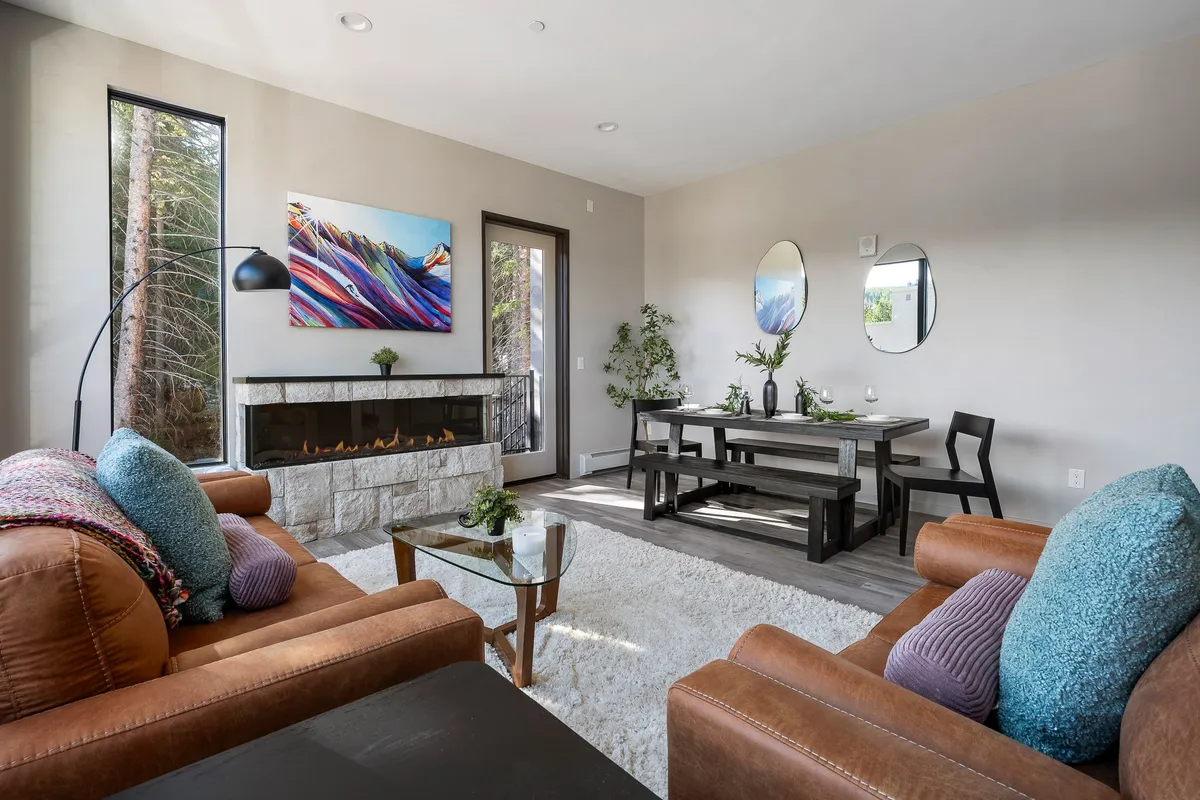 A spacious open-plan living and dining area with a modern fireplace, large windows letting in natural light, colorful wall art, and a dining table. The space is arranged for socializing and relaxing with groups.