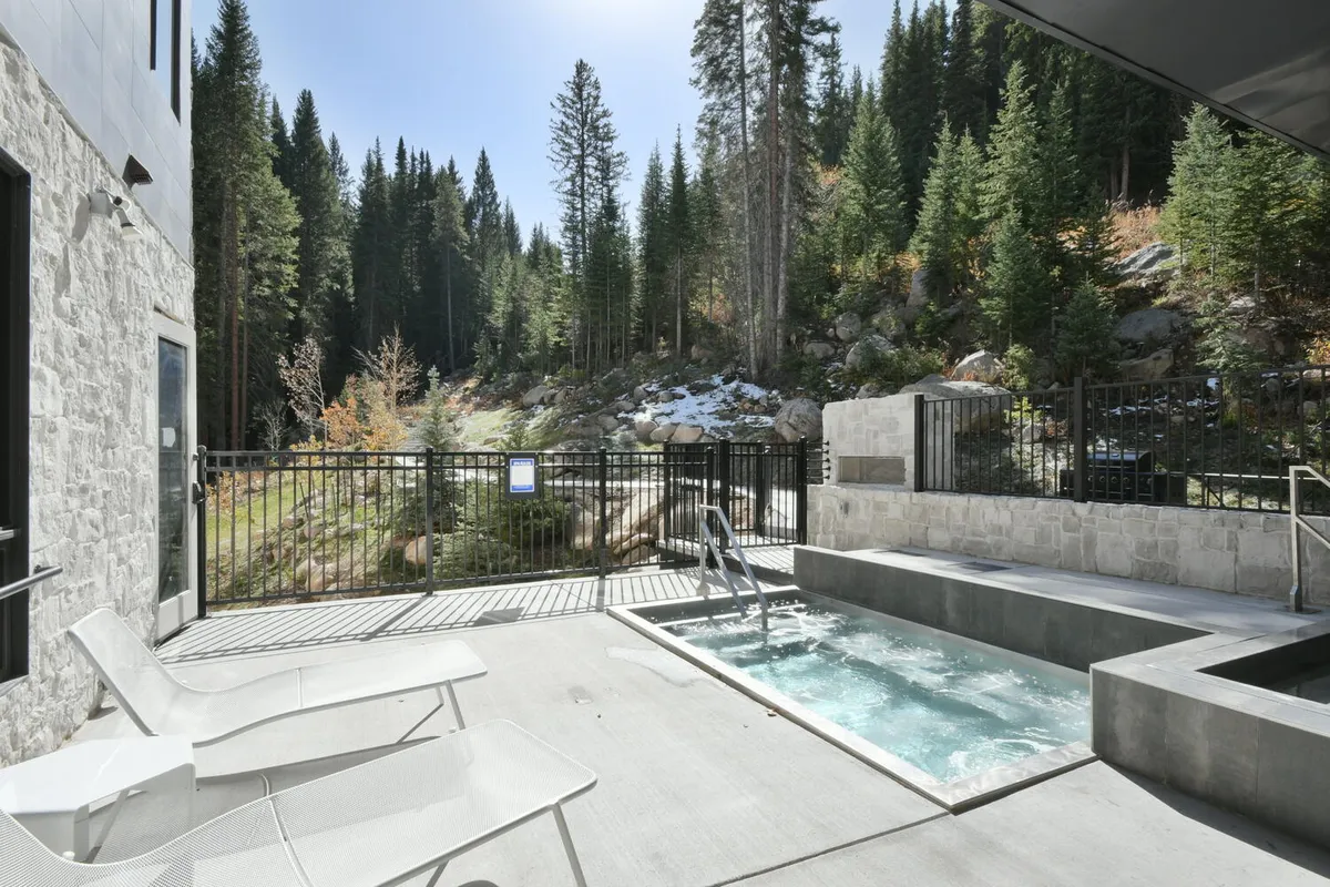 An outdoor hot tub set on a deck surrounded by trees, with deck chairs and a fence framing the serene retreat. The scene is tranquil, yet the setup suggests sociability and post-adventure relaxation, with sunset or stargazing possibilities.