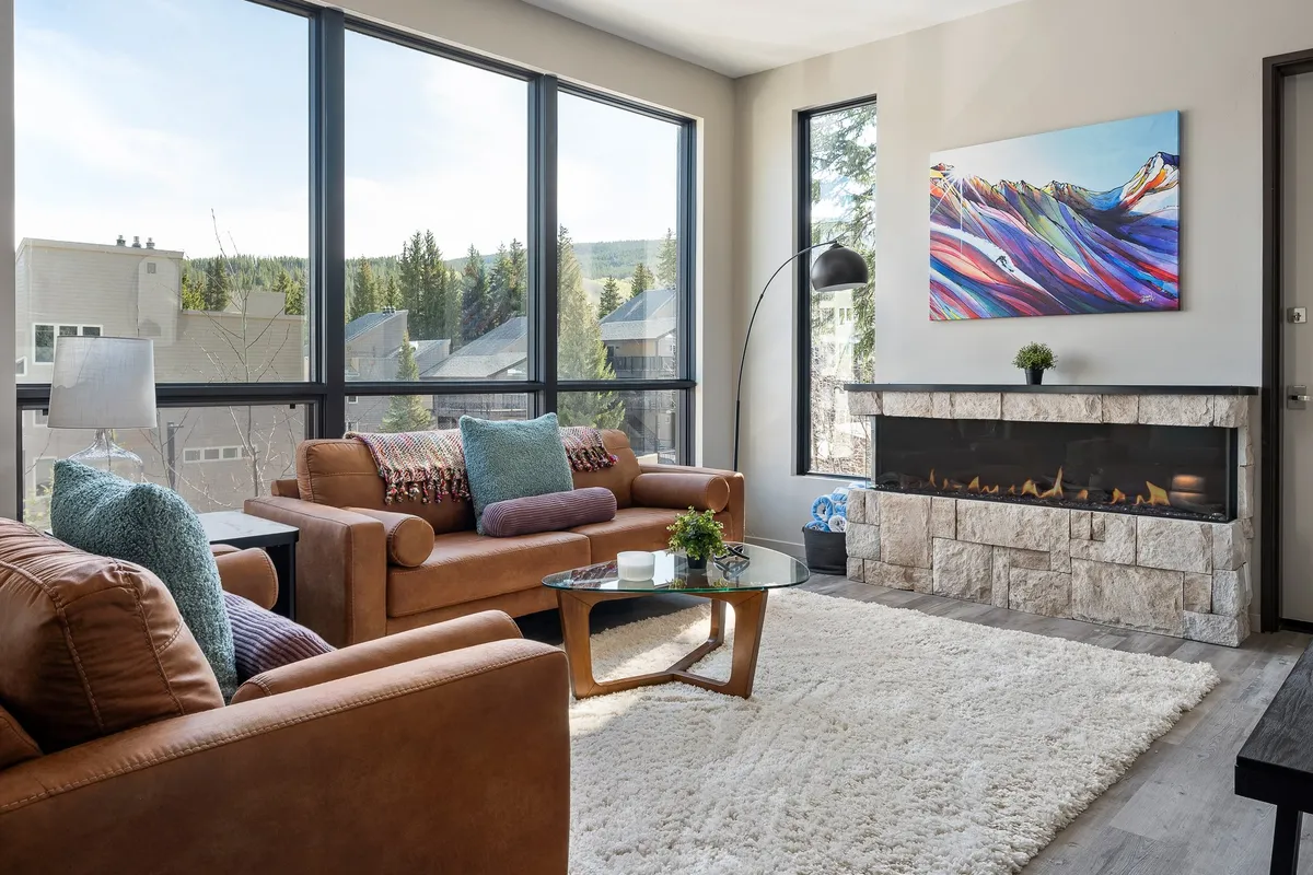 The image features an inviting living area with expansive windows framing breathtaking mountain views, a stylish fireplace at the center, tasteful artwork, and plush, comfortable sofas arranged for socializing and relaxation.