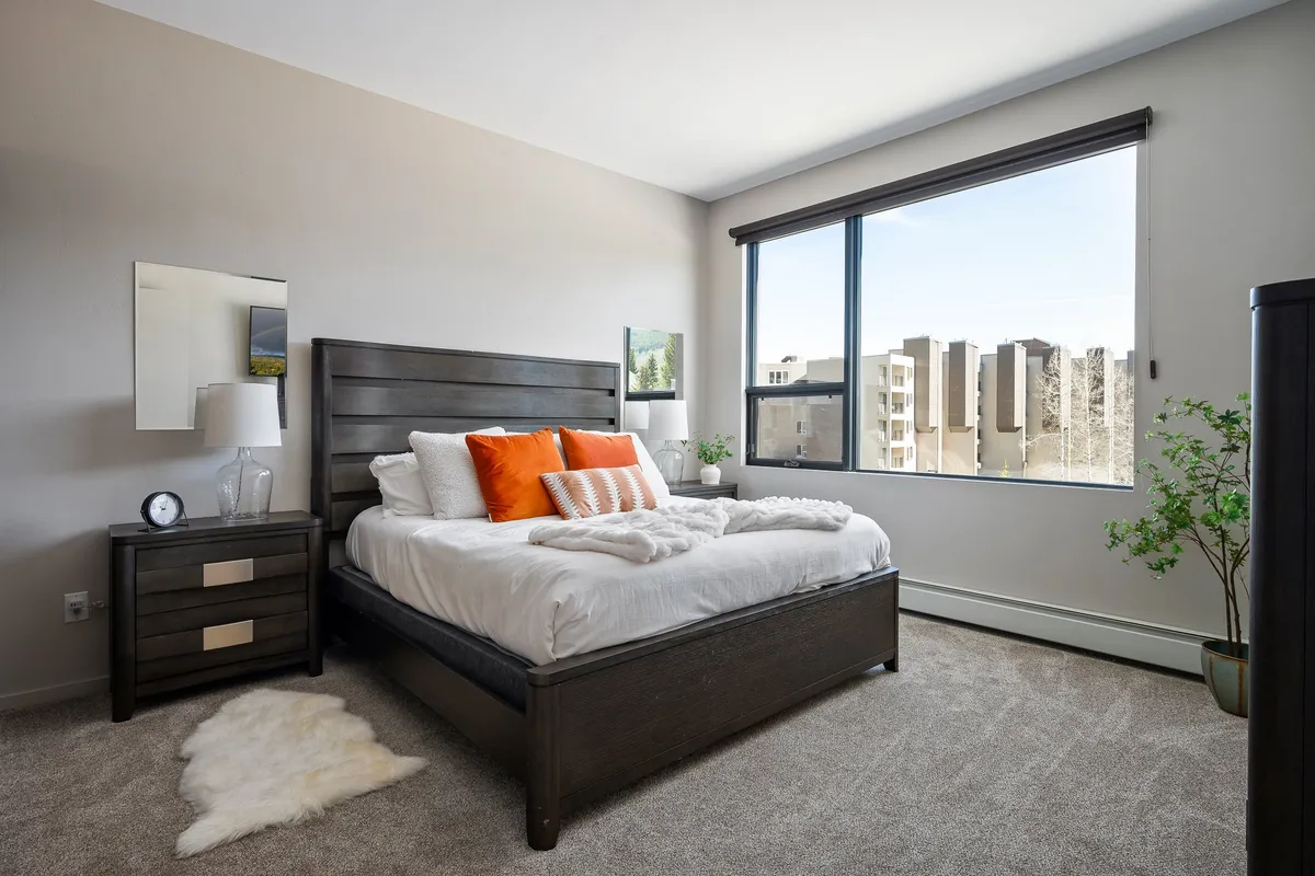 A cozy bedroom featuring a comfortable bed adorned with orange pillows, a large window letting in natural light, and a nightstand with a lamp. The space feels serene and inviting, ideal for restful nights or quiet reading.