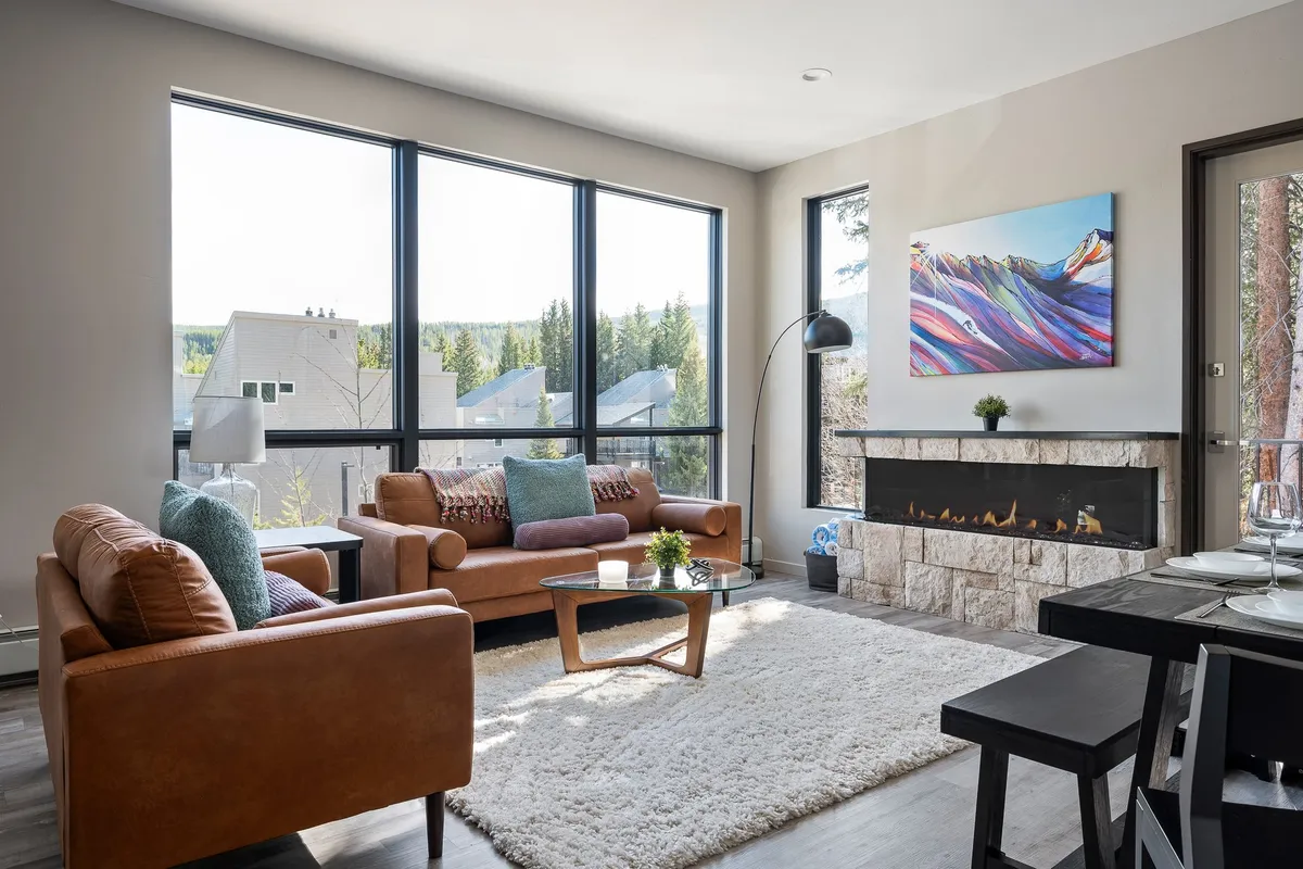 A cozy living area featuring large windows with views of nature, a modern fireplace framed by colorful artwork, and plush, comfortable sofas. The space is bright, with natural light pouring in, and is styled with inviting, contemporary touches that suggest relaxation and togetherness.