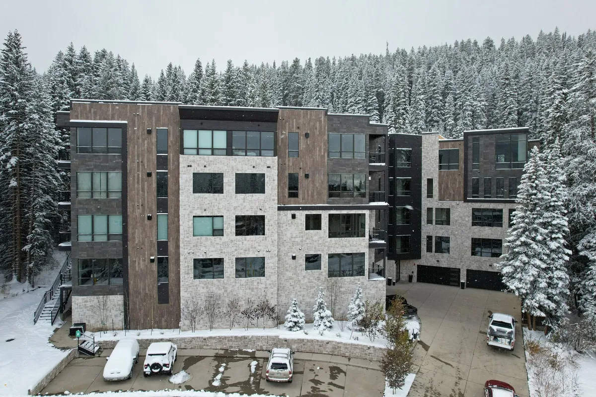 A striking exterior view of a modern building set against a snowy backdrop, surrounded by tall evergreen trees. The inviting architectural design, snow-covered setting, and warm light from inside suggest a cozy retreat after an active day outdoors.