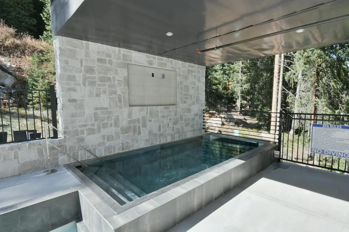 An inviting spa pool area framed by a natural stone wall and surrounded by trees, with a metal railing adding a touch of modernity. The tranquil setting is designed for relaxation and socializing, ideal for enjoying sunset or starlit soaks after mountain adventures.