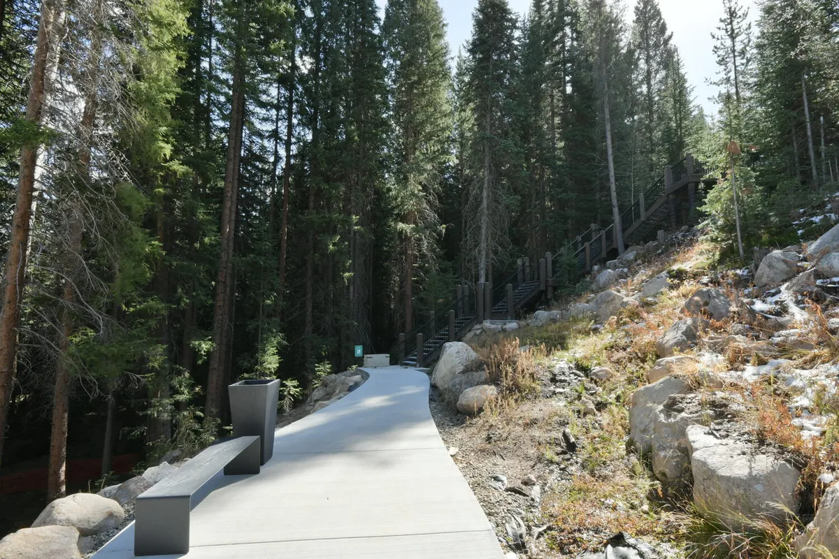 A serene exterior scene featuring a paved walkway winding through lush trees and natural rock formations, with benches placed along the path. The atmosphere is peaceful and inviting, encouraging outdoor exploration and relaxation amidst nature.